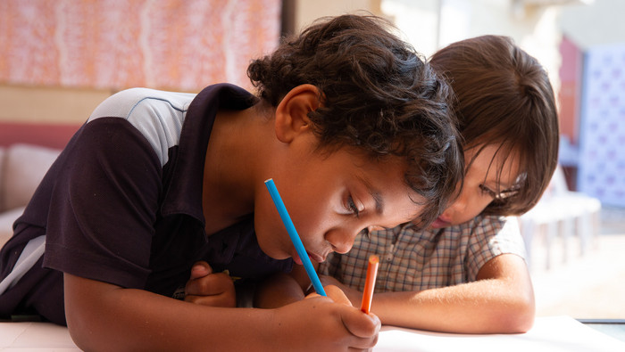 Two children sitting together, focused on drawing and writing at a desk in a learning space.